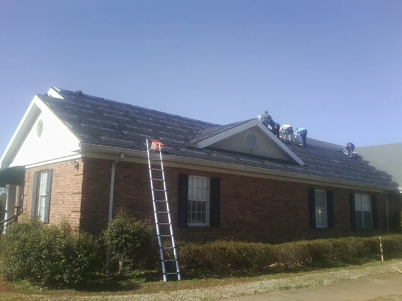 Crew tearing shingles off a brick ranch roof with a ladder set — Metal Roof Repair work by our crew in Kimbolton
