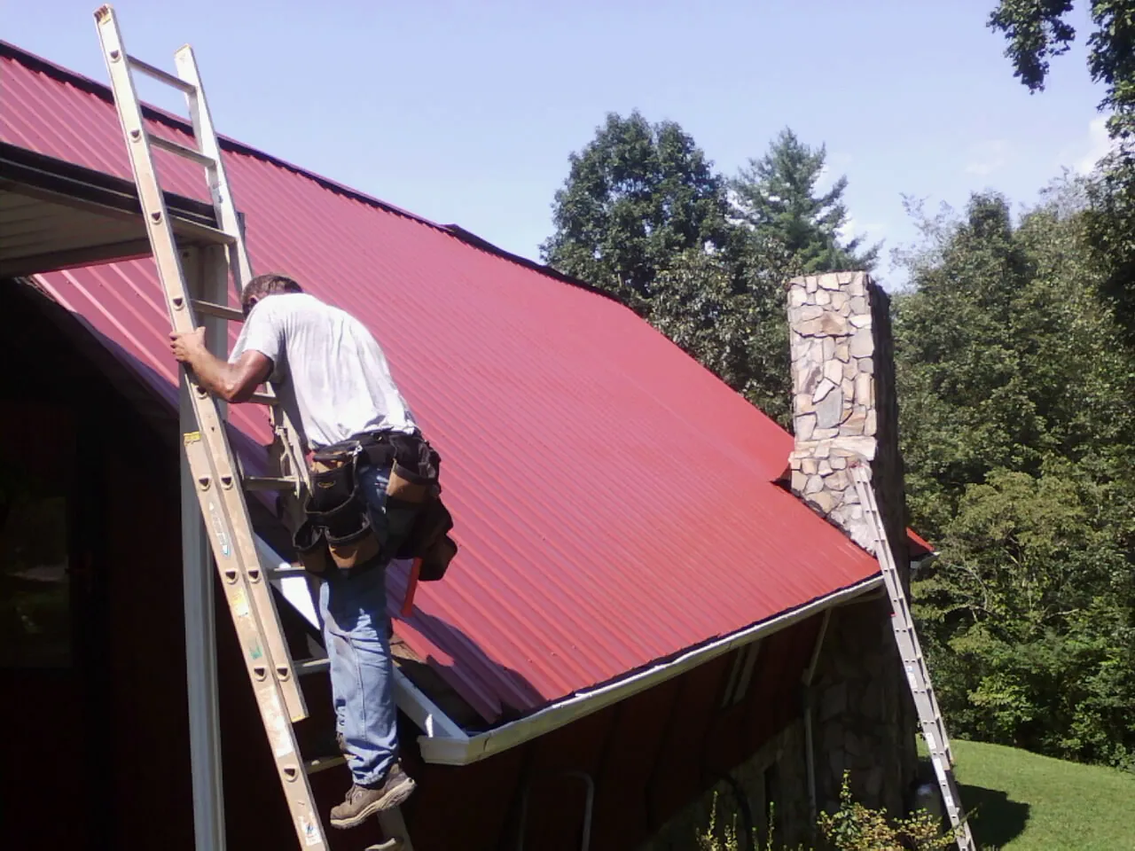 Roofer on a ladder beside a red metal roof and stone chimney — Soffit & Fascia Repair by Roofing Brothers in Kimbolton